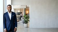Young African American male professional in dark navy blazer and white dress shirt smiling confidently in modern office hallway with blurred colleagues and plants in background.