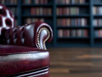Luxurious burgundy leather Chesterfield sofa arm detail with decorative studs against blurred library bookshelves background, creating elegant interior atmosphere.