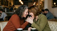Middle-aged man with curly dark hair and woman with shoulder-length brown hair holding hands and laughing together at indoor social gathering, expressing joy and affection.