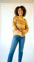 Young woman with curly dark hair and medium skin tone wearing colorful striped knit sweater and blue jeans, smiling at camera against white background for fashion and lifestyle content.