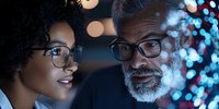 African American woman and mature bearded man with glasses engaged in professional discussion against blurred bokeh lights background. Close-up portrait showing mentorship and teamwork.