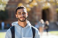Happy young Middle Eastern male college student with backpack smiling outdoors on sunny campus day for education and lifestyle content.