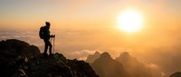 Hiker with trekking poles standing on rocky mountain peak at sunrise, overlooking misty valleys and distant peaks in golden morning light.