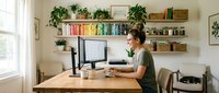 Young man working from home at wooden desk with computer monitor in bright modern office space with floating shelves and green plants.