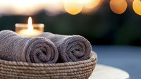 Rolled gray spa towels in woven basket with glowing candle and soft bokeh lights creating relaxing wellness atmosphere for massage and meditation.