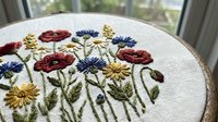 Embroidery hoop with colorful wildflower design featuring red poppies, blue cornflowers and yellow daisies on white fabric for crafting projects.