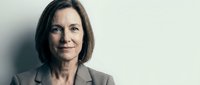 Mature woman with shoulder-length dark hair and fair complexion wearing professional gray blazer, smiling confidently at camera against neutral background. Corporate portrait.