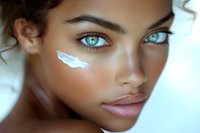 Close up portrait of young mixed race woman with striking blue eyes and glowing skin with white cream streak on cheek against light background.