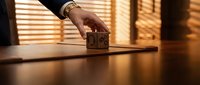 Professional businessman hand placing wooden house model on desk with warm sunlight streaming through blinds creating dramatic shadows for real estate concept.