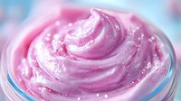 Creamy pink whipped texture with swirls and sparkles in glass container, macro shot showing soft peaks and glossy surface. Cosmetic or dessert background.