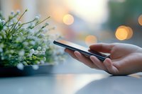 Female hands holding smartphone near window with bokeh lights and white flowers, creating peaceful morning atmosphere. Soft focus lifestyle composition.