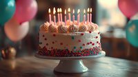 Pink frosted birthday cake with lit candles and colorful sprinkles on white cake stand, surrounded by festive balloons in soft bokeh background for celebration.