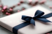 Elegant white gift box wrapped with navy blue satin ribbon bow against blurred festive background with pink bokeh lights, suitable for luxury retail and holiday marketing.