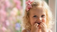 Adorable blonde toddler girl with curly hair and pink bow smiling joyfully while covering mouth with hands in playful gesture against soft floral background.