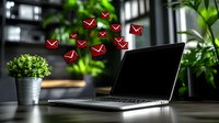 Laptop on desk with floating red email icons, potted plant and green foliage in background. Modern workspace setting with natural light for business communication.