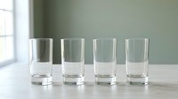 Four empty clear glass tumblers arranged in row on white wooden table near window with natural light and soft green wall background for kitchen decor.