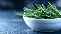 Fresh rosemary sprigs in white ceramic bowl against dark blue textured background, aromatic herbs for cooking and seasoning food preparation.