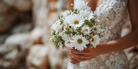 Bride in white lace dress holding fresh daisy bouquet with small white flowers and greenery for wedding ceremony celebration.