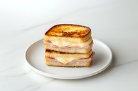 Grilled cheese and ham sandwich stacked on white plate against light background. Melted yellow cheese and pink ham layers between golden toasted bread slices.