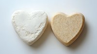 Two heart-shaped bath bombs in white and beige colors on light background, showing natural organic spa cosmetics for aromatherapy and relaxation wellness rituals.