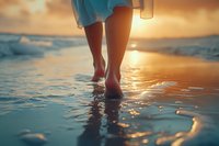 Female legs walking barefoot in shallow ocean water during golden sunset, with waves and sunlight reflection creating serene coastal atmosphere. Perfect for wellness themes.