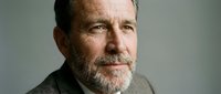 Middle-aged man with gray beard and dark hair wearing neutral blazer against plain background, suitable for corporate, professional, and business applications.