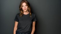 Young Caucasian woman with wavy brown hair wearing dark gray t-shirt against black background, serious expression in professional studio portrait.