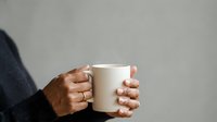 Young adult with dark skin tone holding blank white ceramic mug against neutral gray background, suitable for beverage branding, coffee shop marketing, and product mockup designs.