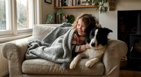 Young woman relaxing with border collie dog on cozy living room sofa wrapped in knit blanket by window creating warm domestic comfort scene.
