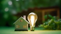 Glowing light bulb next to green house model on natural bokeh background represents eco-friendly home solutions and energy efficiency concepts.