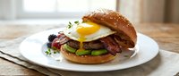 Gourmet bacon cheeseburger with fried egg, avocado and fresh herbs on sesame bun served on white plate. Perfect for restaurant menus and food advertising.