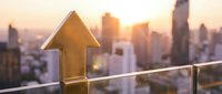 Golden wooden house model on balcony railing with blurred city skyline at sunset, representing real estate investment growth and property market success.