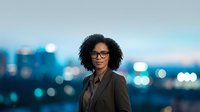 Young woman with curly dark hair and glasses wearing dark blazer, standing against blurred city skyline at dusk. Corporate professional portrait for business, leadership, and career concepts.