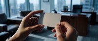 Professional hands holding blank white business card in modern office environment with city skyline view for corporate branding and networking purposes.