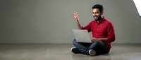 Man with dark hair and beard, appearing to be of Middle Eastern or South Asian descent, wearing a red button-up shirt and jeans, sitting cross-legged on floor while using laptop and gesturing with hand during video call or online meeting.