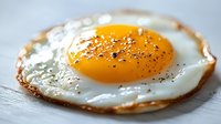 Macro shot of sunny side up fried egg with crispy edges and black pepper seasoning on light wooden surface, showcasing golden yolk and textured whites.