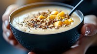 Creamy smoothie bowl topped with mango chunks, chocolate shavings and chia seeds in black ceramic bowl, held by hands. Healthy breakfast concept.