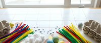 Colorful craft supplies scattered on white tiled floor including wooden sticks, cotton balls, pom poms and paper bags for creative DIY projects and children activities.