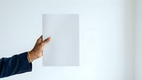 Person with medium skin tone wearing dark navy sleeve holding blank white rectangular card or paper against neutral background for mockup or presentation purposes.