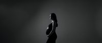 Pregnant woman silhouette in profile against dark background, expecting mother holding belly in dramatic black and white artistic maternity portrait.