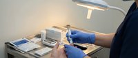 Healthcare professional in blue gloves preparing medical injection syringe at clinical workstation with lamp and supplies for patient treatment procedure.
