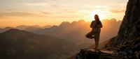 Woman practicing yoga tree pose on mountain cliff at golden sunrise with dramatic alpine landscape and layered peaks in background.