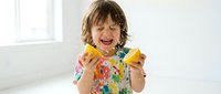 Happy young child with shoulder-length brown hair and fair complexion, wearing colorful floral shirt, holding and eating fresh mango slices in bright, white studio setting.