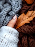 Cozy autumn composition with female hand in white knitted sweater touching soft gray and brown yarn balls with dried orange oak leaf, creating seasonal mood.