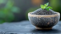 Black chia seeds in rustic ceramic bowl with fresh mint leaf garnish on dark stone surface against blurred green background, macro food photography.