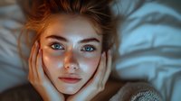 Young woman with bright blue eyes and glowing skin lying on white bedding, hands framing face, captured in moody teal lighting with soft focus effect.