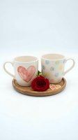 Pair of ceramic mugs with heart and polka dot designs on wooden tray with red rose and love tag for Valentine's Day gift or romantic home decor.