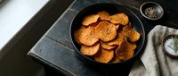 Crispy baked sweet potato chips in a dark bowl on a rustic wooden table with herbs and spices, healthy snack concept.