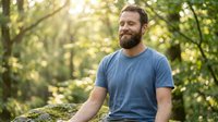 Middle-aged Caucasian man with full beard wearing blue t-shirt standing in sunlit forest surrounded by green foliage and moss-covered rocks, outdoor portrait for lifestyle and nature themes.