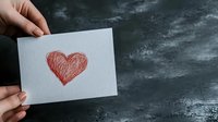 Red heart drawn on white paper held against dramatic stormy sky background, representing hope, love and resilience during dark times.